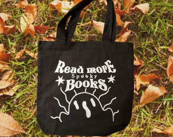 Puede incluir: Bolsa de lona negra con texto blanco que dice "Read more Spooky Books" y un sol estilizado con una cara de fantasma.