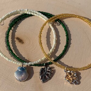 May include: Three beaded bracelets with charms. The bracelets are made with white, green, and gold beads. The charms are a seashell, a monstera leaf, and a starfish.