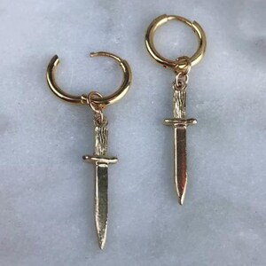 May include: A pair of gold-toned hoop earrings, each featuring a dangling charm shaped like a small dagger. The dagger charms have a textured handle and a smooth, pointed blade. The hoops are circular and polished, providing a metallic finish.