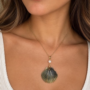 May include: A gold chain necklace featuring a pendant with a single white pearl and a dark green, ribbed seashell. The shell is secured with gold wire wrapping, and the necklace rests against the neck, showcasing a delicate and refined design.
