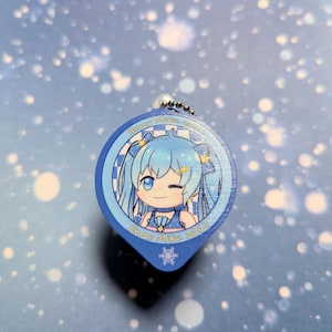 May include: A blue and white Snow Miku 2017 charm. It features a cartoon character with blue hair and a winking expression. The charm has a snowflake design and a silver chain for attachment.