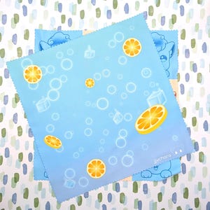 May include: A square light blue microfibre cleaning cloth with a scalloped edge, featuring a pattern of orange slices, ice cubes, and white bubbles. The text "jychara" is printed in the bottom right corner with a small cat-like icon.