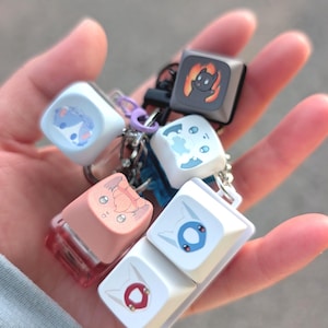 Puede incluir: Una colección de seis teclas mecánicas cuadradas para teclado, unidas a un llavero de metal. Las teclas presentan varias ilustraciones de personajes de dibujos animados en colores blanco, azul claro, melocotón y gris oscuro con fuego.