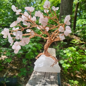 May include: A pink rose quartz wire wrapped tree with a white quartz base. The tree is on a wooden surface with green foliage in the background.