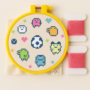 Può includere: Un telaio da ricamo a punto croce con una cornice gialla, con personaggi di cartoni animati pixelati in vari colori. Il design include un pallone da calcio, cuori e stelle. Due rocchetti di filo rosa sono accanto al telaio. Il testo "cogg art" è in basso a destra.