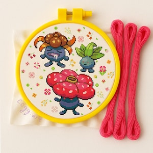 Peut inclure: Un cercle de broderie jaune présente une broderie au point de croix avec trois personnages de dessins animés et des motifs floraux. Le tissu crème porte la mention "cogg. art". Trois brins de fil à broder rose vif sont à droite.