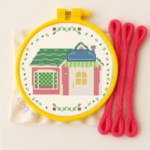 Peut inclure: Un kit de point de croix représentant une maison rose avec un toit vert et une fenêtre jaune. Le motif est encadré dans un cercle à broder jaune. Trois brins de fil à broder rose vif sont à côté du cercle. Le kit comprend un petit tissu beige avec le texte "cogg. art".
