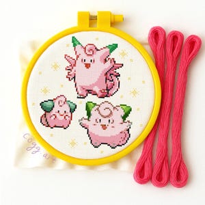 Può includere: Un telaio da ricamo a punto croce con tre personaggi dei cartoni animati rosa con dettagli verdi, su sfondo bianco con stelle gialle. Il telaio giallo è accompagnato da tre fili di filo da ricamo rosa.