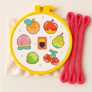 May include: A yellow embroidery hoop holds a cross-stitch design of various fruits, including an orange, apple, peach, pear, lime, and cherries. The design is surrounded by a yellow hoop, with three strands of bright pink embroidery floss to the right. The text "cogg art" is visible.