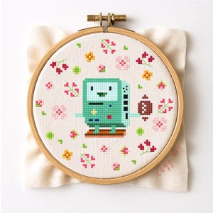 Op de afbeelding: Een kruissteek borduurring met een gepixeleerd groen personage en een bruin object, omringd door roze, gele en rode bloemmotieven. Het ontwerp staat op crèmekleurige stof, gehouden in een houten ring. De tekst "cozy art" is zichtbaar.