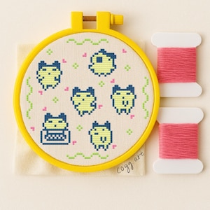 Può includere: Un telaio da ricamo a punto croce con personaggi pixelati gialli e blu su tessuto color crema. Il telaio giallo tiene il disegno, con rocchetti di filo rosa a destra. Il disegno include il testo "cogg art".