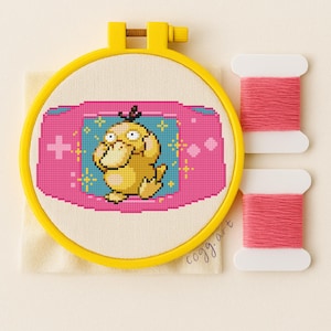 May include: A cross-stitch embroidery featuring a yellow Psyduck character within a pink and blue game console design, set in a yellow hoop. Two spools of pink thread are next to the hoop. The artist's signature "cogg.art" is visible.