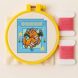 May include: A cross-stitch embroidery featuring an orange dragon design within a blue pixelated frame, set in a yellow embroidery hoop. Two spools of pink thread are positioned to the right. The design includes stars and flowers.