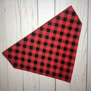 May include: Red and black buffalo plaid dog bandana. The bandana is a triangle shape with a classic check pattern. The fabric is a medium weight and appears to be cotton or a cotton blend. The bandana is laying on a light wood surface.