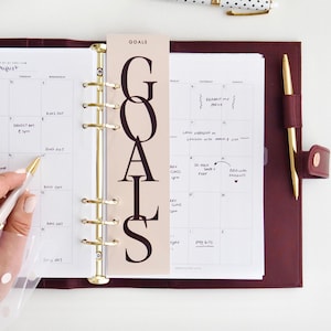 May include: A burgundy leather planner with gold accents, open to a calendar view. A "GOALS" bookmark is inserted. A polka-dot pen and gold paper clips are on the white surface. A hand is holding a gold pen.
