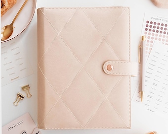 May include: A pale pink quilted planner with a button clasp, alongside gold stationery items, a pen, and a card reading "today". The planner's diamond pattern and soft colour scheme create a stylish aesthetic. The scene includes a croissant.