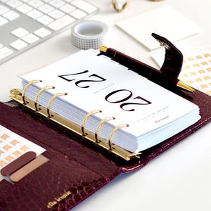 May include: A deep purple, crocodile-textured leather personal planner with gold-toned ring binding and a matching pen. The open planner displays a white page with the text 20 27 and the brand name ella iconic printed in black ink.