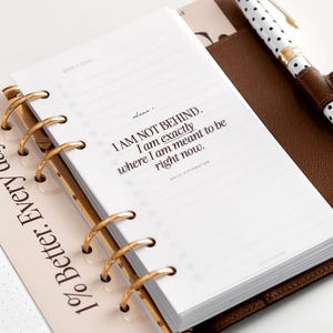 Pode incluir: Agenda aberta com argolas douradas, apresentando uma página branca com o texto "I AM NOT BEHIND. I am exactly where I am meant to be right now." Uma caneta com corpo branco e bolinhas pretas está no bolso da agenda.