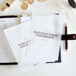 May include: A flat lay arrangement with a planner, pen, gold earrings, bracelet, sunglasses, and makeup. The planner pages display the text "WHAT'S MEANT FOR ME it won't pass me by" and "IT WILL at the ri".