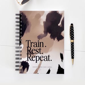 Può includere: Un diario di fitness rilegato a spirale con le parole "Train. Rest. Repeat." in nero. La copertina presenta un design astratto beige e nero. Una penna a pois bianchi e neri e occhiali da sole sono anche nell'immagine.