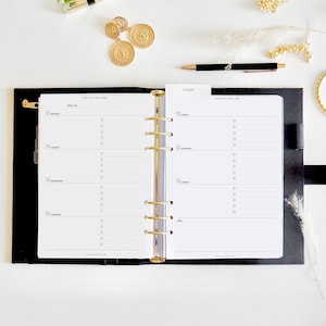 May include: Black leather planner with gold detailing, open to a weekly spread. The planner has a gold binder and is surrounded by a black pen, gold coins, and a gold lipstick on a white surface.
