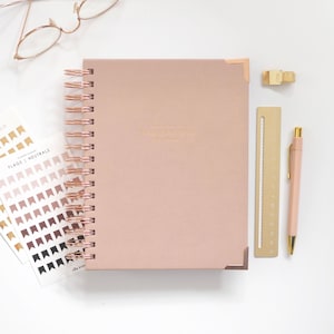 May include: A dusty rose spiral-bound planner with rose gold metal corner protectors and a matching coil. Beside it are rose gold glasses, a sheet of flag-shaped stickers labeled FLAGS | NEUTRALS, a gold metal ruler, a gold clip, and a rose gold pen.
