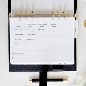 May include: A black leather planner with gold rings and a Social Media Content Planner insert. The planner is open, revealing the insert with handwritten notes and a pen. The insert includes sections for Instagram, TikTok, Pinterest, and Facebook.