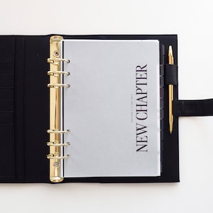 May include: Black leather binder with gold detailing, open to a page with the words "NEW CHAPTER". A gold pen is held in a black loop. The binder is on a white surface.