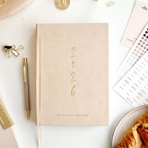 Può includere: Un'agenda beige del 2026 con scritte dorate e una penna abbinata. L'agenda è etichettata "CEO OF MY OWN LIFE PLANNER". Accessori dorati, tra cui orecchini, fermagli e un piccolo oggetto geometrico, sono sparsi.