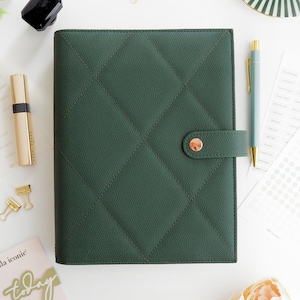 May include: A dark green, quilted planner with a rose gold clasp and matching pen. The planner is surrounded by stationery items, including a gold pen, gold paper clips, and a small card that says "today".