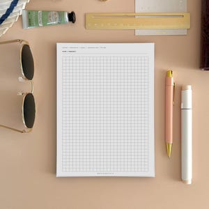 May include: A white grid notepad with the text NOTES / PROJECTS / IDEAS / JOURNALING / TO DO and DATE / SUBJECT at the top. The notepad is surrounded by a pair of sunglasses, a gold ruler, a peach pen, a white marker, two gold hoop earrings, and a tube of cream.