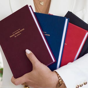 May include: A collection of four hardback planners in burgundy, navy blue, red, and black. Each planner features gold foil text on the front cover that reads twenty twenty seven life planner. The books have white page edges and a smooth matte finish.