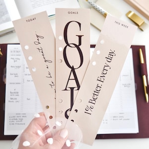 Può includere: Tre inserti per planner beige con testo: "Today to be a great day", "GOALS" e "This Week 1% Better. Every day." Gli inserti hanno un bordo perforato e sono tenuti davanti a un planner.