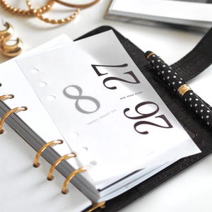 Peut inclure: Un classeur en cuir noir avec des anneaux dorés contenant des pages de planning blanches. La page supérieure affiche le texte 8 27, les mots MID YEAR RESET et TWENTY TWENTY SIX. Un stylo noir à pois blancs repose sur le classeur près de créoles dorées.