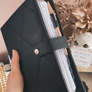Op de afbeelding: Zwarte leren planner met een ruitpatroon en een gouden sluiting. De planner is open en toont gelabelde verdelers en een roségouden pen. Ontworpen voor organisatie en planning.