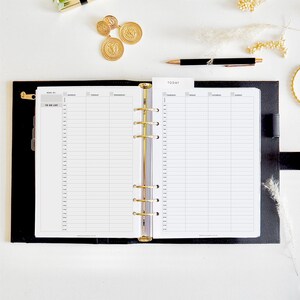 Könnte beinhalten: Ein offener schwarzer Planer mit goldenen Akzenten, der ein tägliches Zeitplan-Layout zeigt. Der Planer ist von goldenen Accessoires umgeben, darunter Münzen, ein Lippenstift und ein Stift, die alle auf einer weißen Oberfläche angeordnet sind. Der Planer hat einen 'Heute'-Reiter.