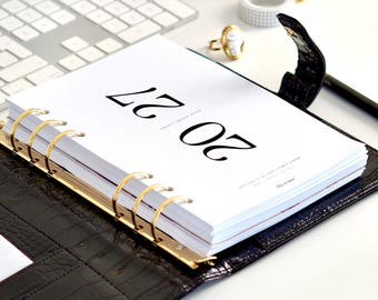 May include: A black crocodile-textured leather planner with gold-toned rings, featuring a white title page with the text 20 27 printed in large black font. The planner is open, displaying white pages, and includes the text ella iconic in gold.