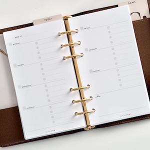 May include: An open planner with gold rings and white pages. The planner displays a weekly spread with sections for each day, including "Today" and "Goals" tabs. The planner is in a brown leather case. The brand name "ELLA ICONIC" is visible.