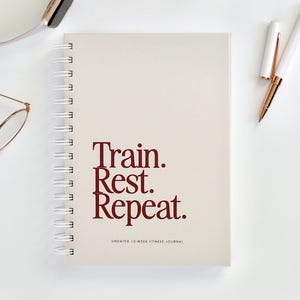 Op de afbeelding: Crèmekleurig spiraalgebonden fitnessdagboek met de woorden "Train. Rest. Repeat." in bordeauxrood. Het dagboek is een ongedateerd 12-weeks fitnessdagboek. Een smartwatch, bril, pen en oorbellen zijn ook zichtbaar.