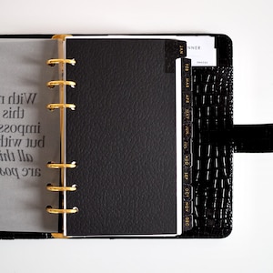 Pode incluir: Uma agenda de argolas preta com textura de crocodilo, apresentando argolas douradas e divisórias mensais rotuladas de JAN a DEC em letras douradas. Uma página de papel vegetal translúcido com texto impresso é visível à esquerda.