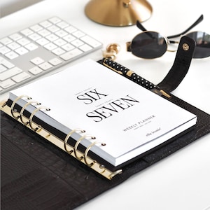 May include: A black leather weekly planner with gold rings and accents, open to a page reading "SIX SEVEN." A polka-dot pen, sunglasses, and a keyboard are also visible on the white surface.
