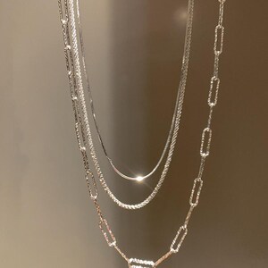May include: Three silver necklaces of different styles hang against a neutral backdrop. The necklaces include a fine chain, a textured rope chain, and a chain with elongated oval links. The chains are all silver in colour.