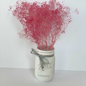 May include: A white painted mason jar with a gray ribbon and the text "Ball Mason" on the front. The jar is filled with a bouquet of pink dried baby's breath.