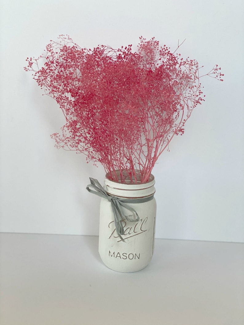 May include: A white painted mason jar with a gray ribbon and the text "Ball Mason" on the front. The jar is filled with a bouquet of pink dried baby's breath.