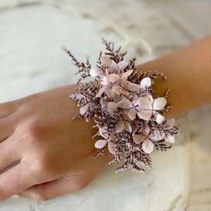 May include: A delicate wrist corsage made with pink and white dried flowers. The flowers are arranged in a cluster and tied with a thin white ribbon.
