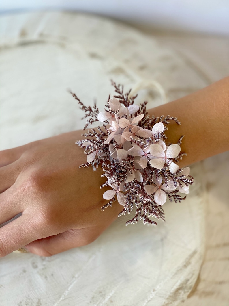 May include: A delicate wrist corsage made with pink and white dried flowers. The flowers are arranged in a cluster and tied with a thin white ribbon.