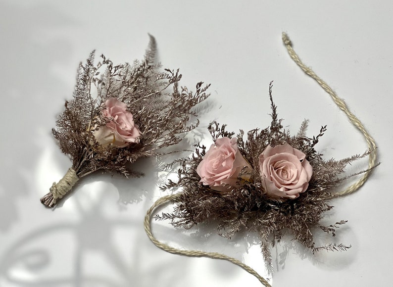 May include: Two small bouquets of dried flowers with pink roses and brown dried grasses. The bouquets are tied with twine and are connected by a long piece of twine.