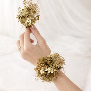 以下が含まれることがあります： 金色の花のリストコサージュと、お揃いのブートニア。 リストコサージュは白いリボンで固定されています。 ブートニアは手で持っています。 花は金属仕上げで、結婚式やフォーマルなイベントに最適です。