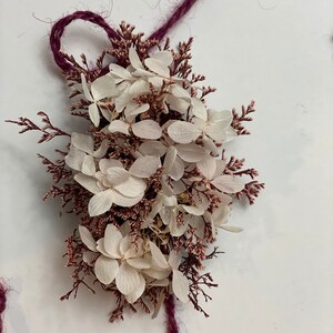 May include: A bouquet of dried white hydrangea flowers with pink stems tied together with a burgundy twine.