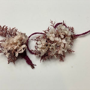 May include: Two small bouquets of dried flowers tied with burgundy twine. The bouquets are made of white hydrangeas and pink sprigs.
